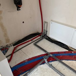 HYDROLINE SPÓŁKA Z OGRANICZONĄ ODPOWIEDZIALNOŚCIĄ - Instalacja ogrzewania podłogowego w trakcie realizacji: czerwone i niebieskie rury ułożone na izolacji, szare rury odpływowe, białe płyty maskujące na ścianie.