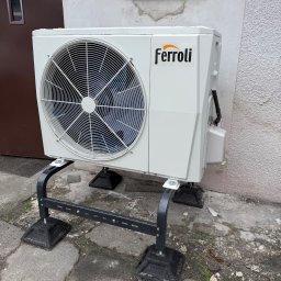 HYDROLINE SPÓŁKA Z OGRANICZONĄ ODPOWIEDZIALNOŚCIĄ - Biała pompa ciepła Ferroli zamontowana na czarnej, metalowej konstrukcji, ustawiona na betonowym podłożu przy ścianie budynku z oknem.