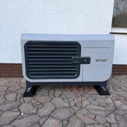 HYDROLINE SPÓŁKA Z OGRANICZONĄ ODPOWIEDZIALNOŚCIĄ - Szara pompa ciepła Airmax³ na kostce brukowej, ustawiona przy białej ścianie budynku z widocznym oknem. Widoczne detale konstrukcyjne i logo producenta.