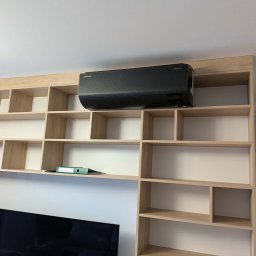 HYDROLINE SPÓŁKA Z OGRANICZONĄ ODPOWIEDZIALNOŚCIĄ - Czarna jednostka klimatyzacji Samsung WindFree zamontowana nad jasnym, drewnianym regałem z białymi plecami, częściowo zakrywającym telewizor.