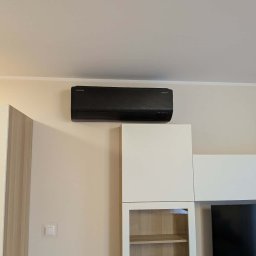 HYDROLINE SPÓŁKA Z OGRANICZONĄ ODPOWIEDZIALNOŚCIĄ - Czarny klimatyzator Samsung WindFree zamontowany wysoko na jasnej ścianie w nowoczesnym salonie, nad białymi meblami modułowymi.