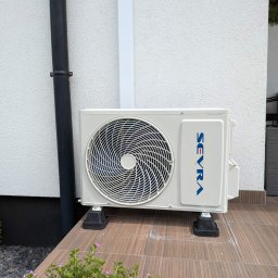 HYDROLINE SPÓŁKA Z OGRANICZONĄ ODPOWIEDZIALNOŚCIĄ - Zewnętrzna jednostka klimatyzatora SEVRA na czarnych podstawkach, stojąca na brązowych płytkach tarasu, obok białej, teksturowanej ściany budynku i czarnej rynny.