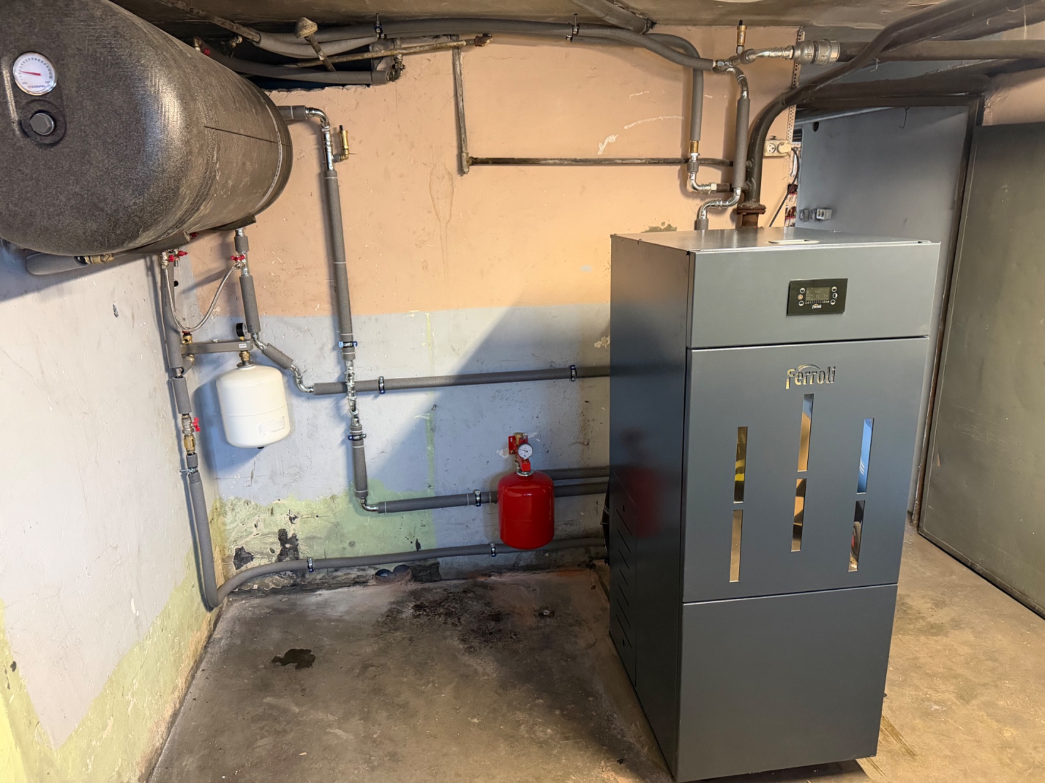 Szary piec Ferroli z podłączonymi instalacjami: zbiornik, rury w izolacji, naczynie przeponowe. Widoczne elementy instalacji grzewczej w piwnicy.