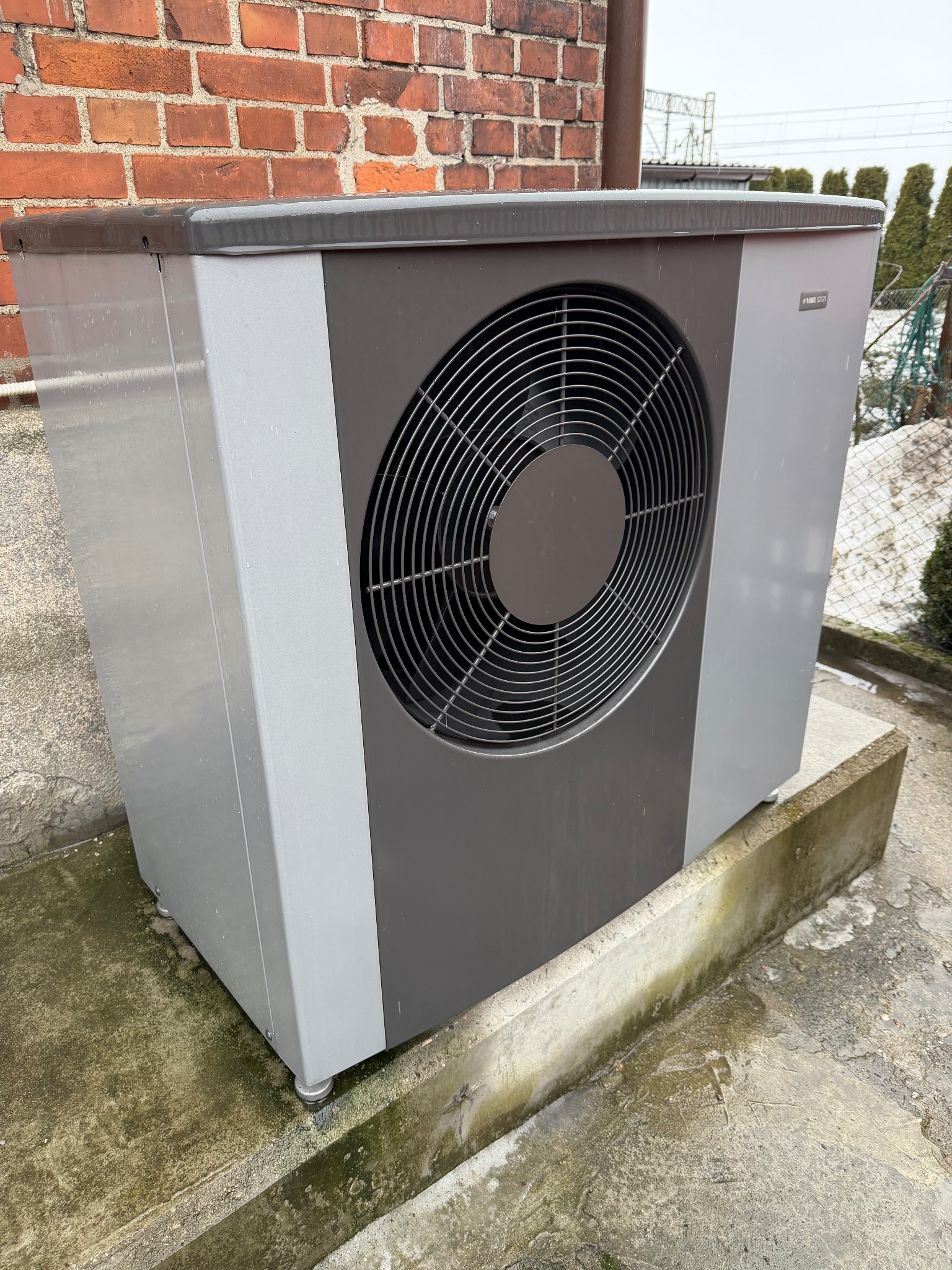 Szaro-grafitowa pompa ciepła na betonowym fundamencie, z widoczną ceglaną ścianą w tle i elementami zieleni. Widoczne ślady wilgoci na urządzeniu i podłożu.