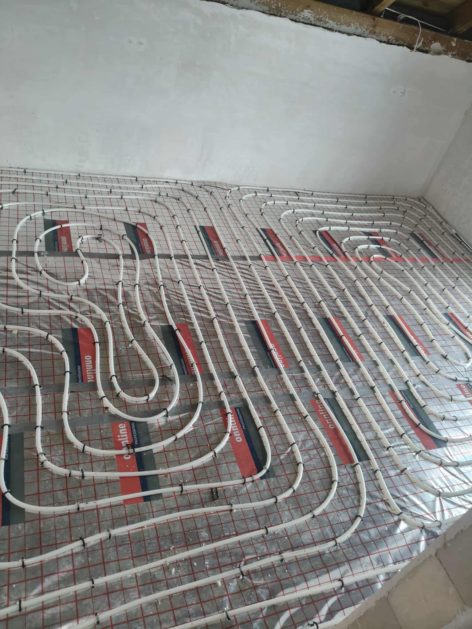 Białe rury ogrzewania podłogowego ułożone na folii z czerwoną siatką, widok z góry, surowy stan wykończenia wnętrza, przygotowanie do wylewki.