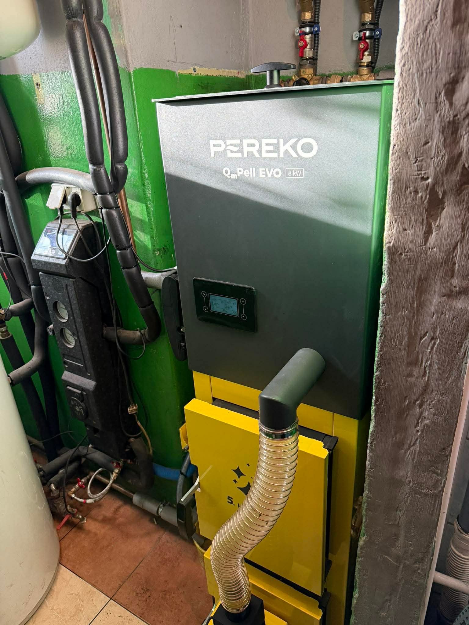 Szaro-żółty piec Pereko Qm Pell EVO 8kW w kotłowni, widoczne rury instalacji grzewczej i sterownik. Ściany pomalowane na zielono, podłoga wyłożona płytkami.
