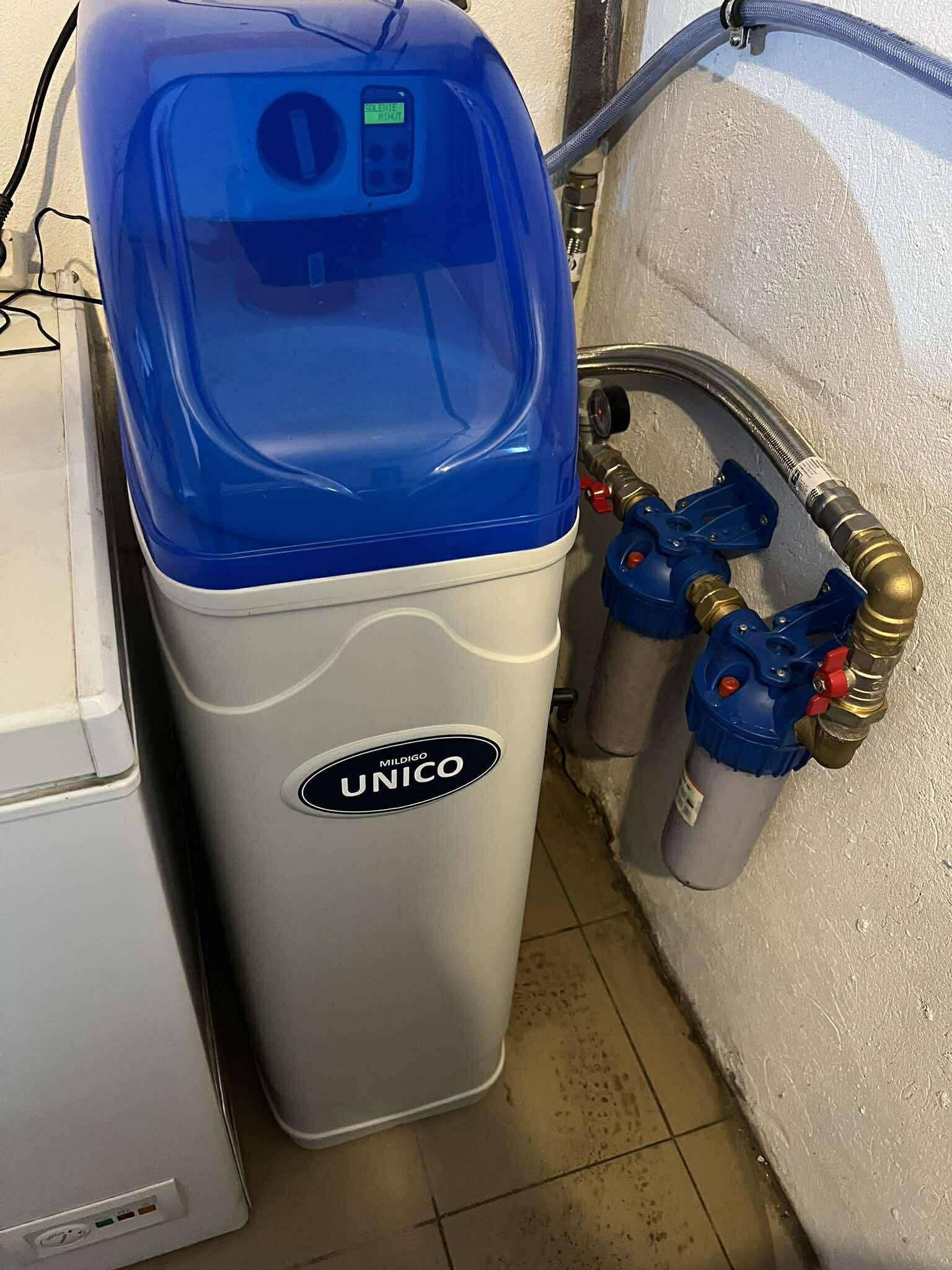 Zamontowany zmiękczacz wody Mildigo Unico z podwójnym filtrem mechanicznym, widoczne przyłącza hydrauliczne i wyświetlacz. Urządzenie stoi obok zamrażarki.