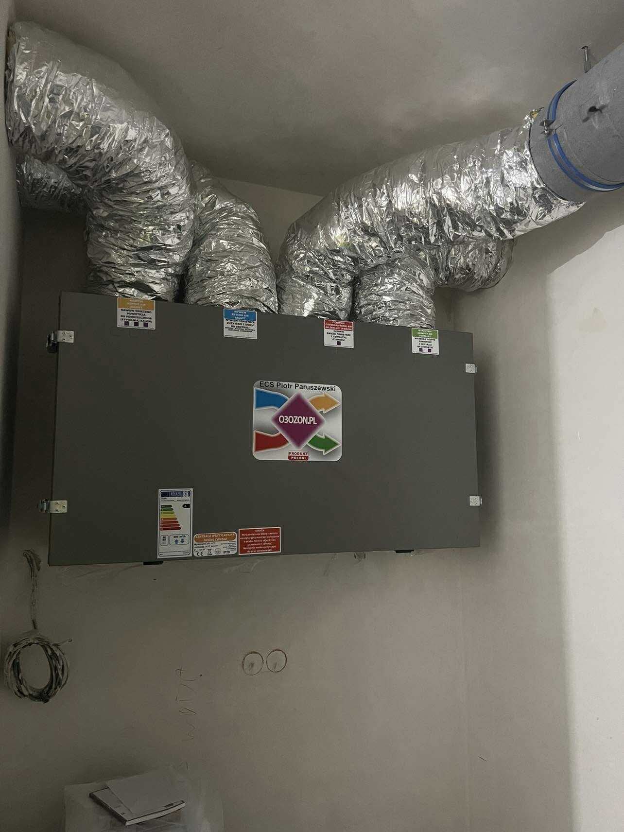 Szara centrala wentylacyjna z podłączonymi, elastycznymi przewodami wentylacyjnymi w folii aluminiowej, zamontowana w pomieszczeniu. Widoczne naklejki informacyjne i logo producenta.