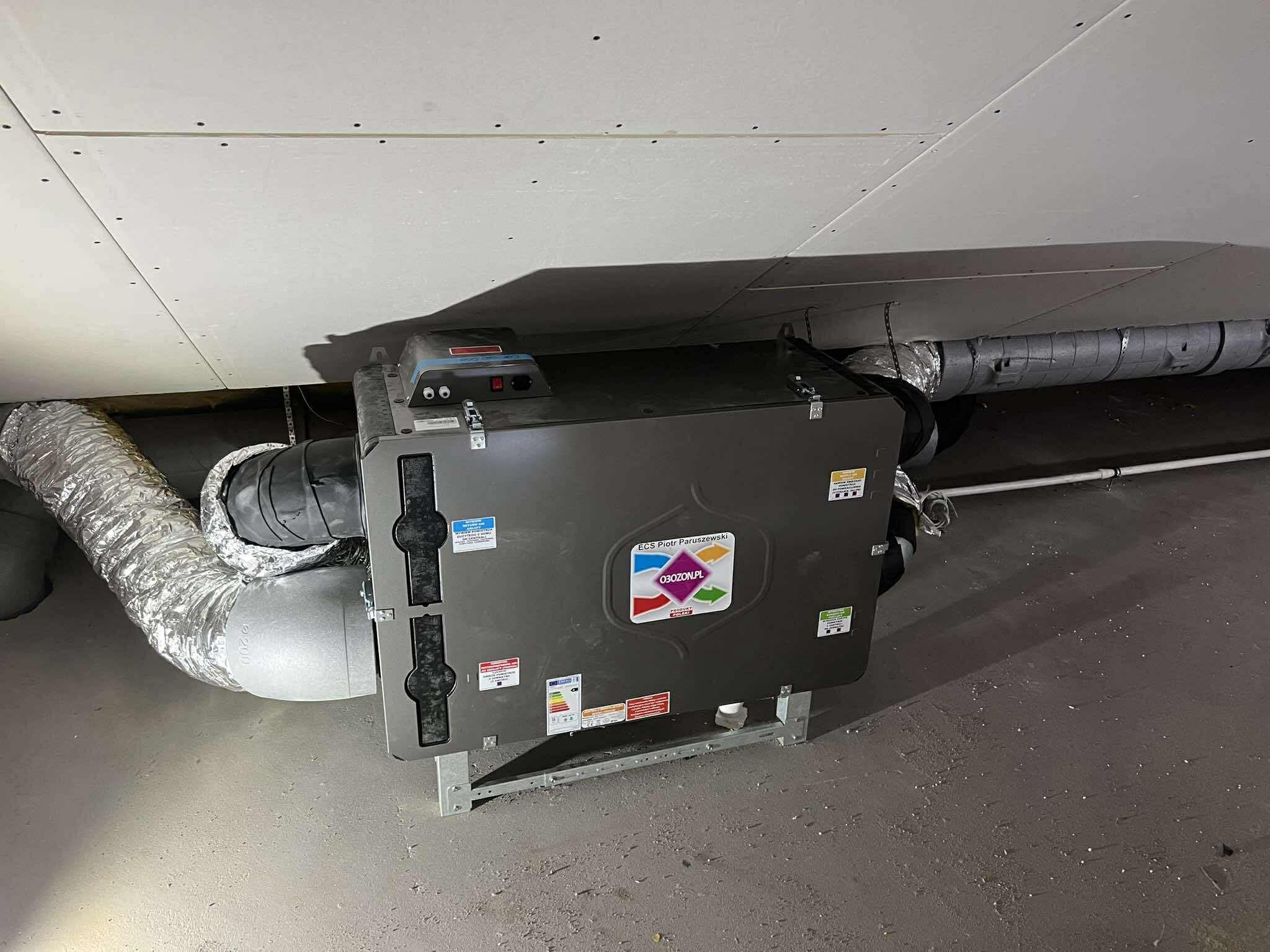 Szara centrala wentylacyjna z podłączonymi przewodami wentylacyjnymi, zamontowana na poddaszu z płyt gipsowo-kartonowych. Widoczne elementy montażowe i etykiety informacyjne.