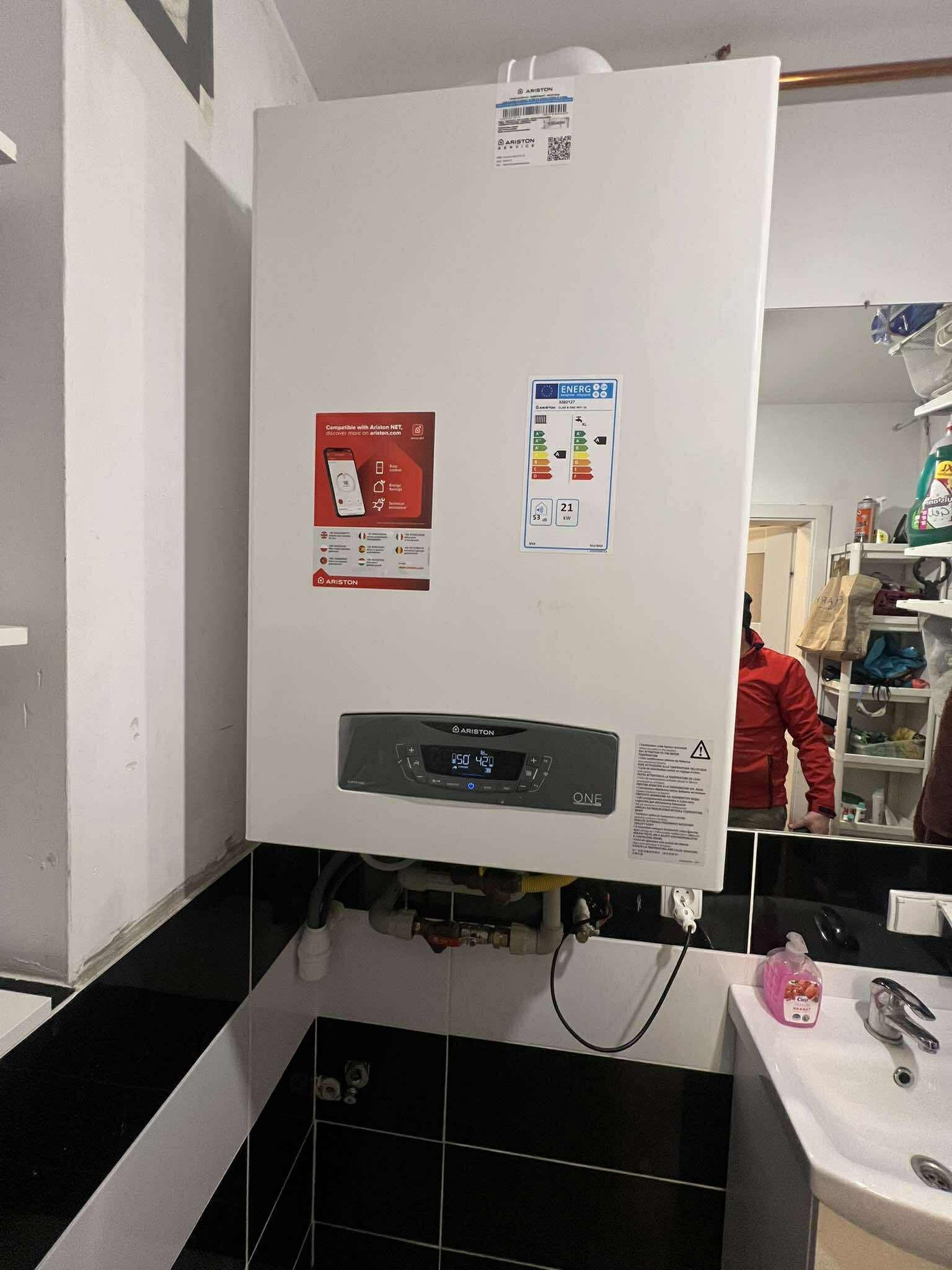 Biały, wiszący piec gazowy Ariston ONE w łazience z czarno-białymi płytkami. Widoczne naklejki informacyjne i podłączenia hydrauliczne. Odbicie osoby w lustrze.