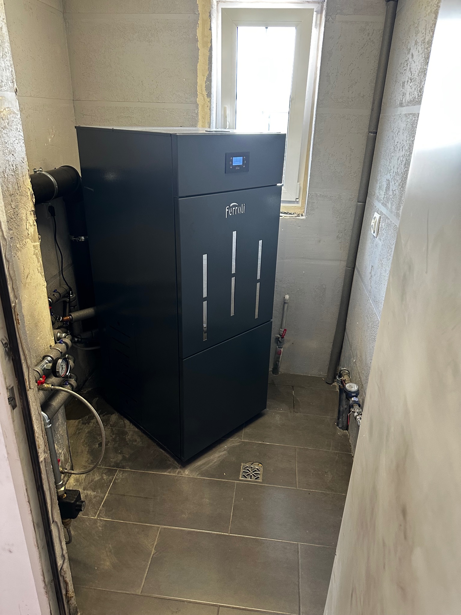 Czarny piec Ferroli w kotłowni z szarymi płytkami i instalacją hydrauliczną. Widoczne rury, zawory i okno. Ściany surowe, w trakcie wykończenia.