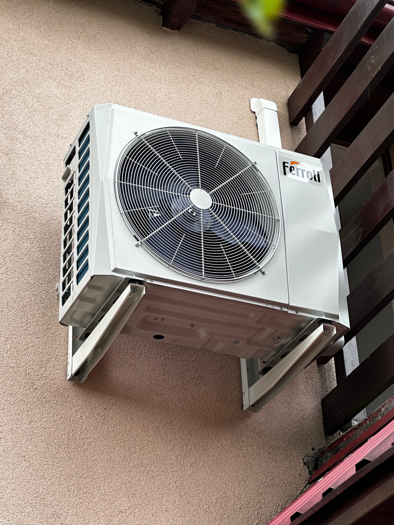 Biała jednostka zewnętrzna klimatyzacji Ferroli zamontowana na elewacji budynku, widoczna z dołu pod kątem, z fragmentem drewnianej konstrukcji w tle.