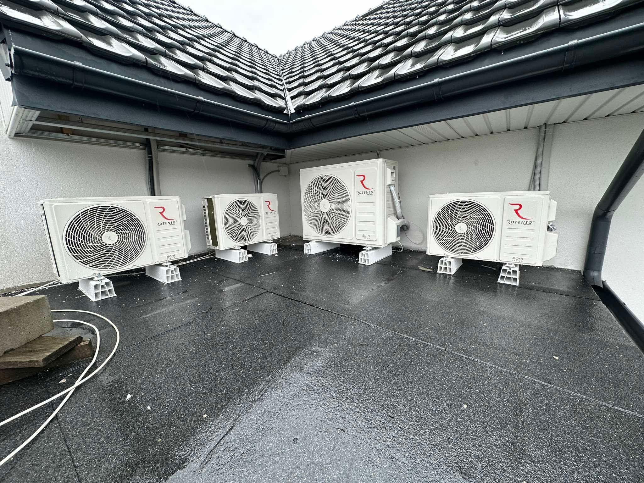 Cztery klimatyzatory Rotenso ELIS Series zamontowane na dachu, każdy na betonowych podstawach. Widoczne przewody i fragment dachu z dachówką. Dach pokryty papą.