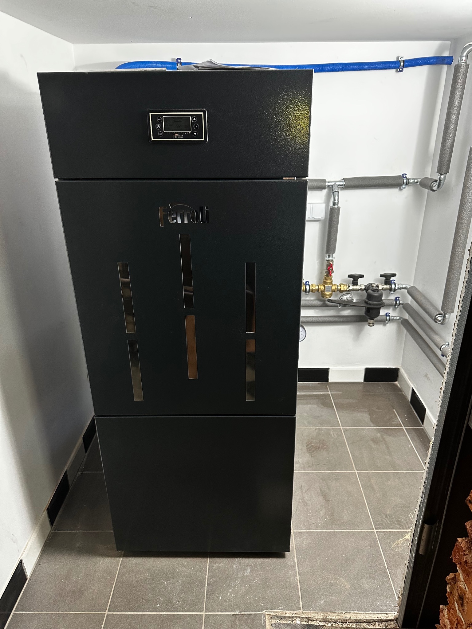 Czarny piec Ferroli z widocznym panelem sterowania i instalacją hydrauliczną w tle, zamontowany w pomieszczeniu z szarą podłogą. Ujęcie z niskiej perspektywy.