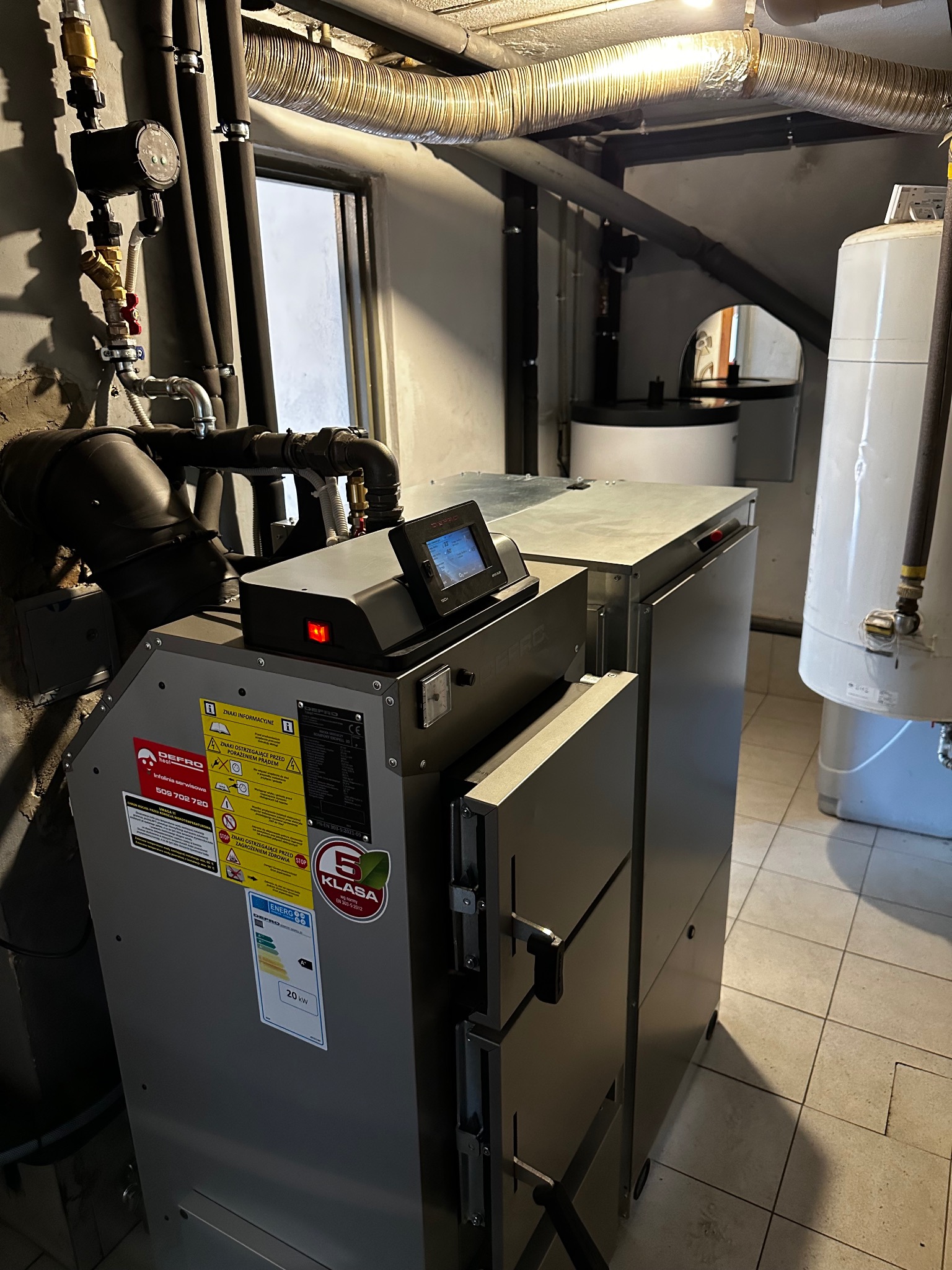 Nowoczesny piec na paliwo stałe Defro z panelem sterowania, widoczne rury instalacji grzewczej i zasobnik ciepłej wody w kotłowni. Klasa energetyczna A+.