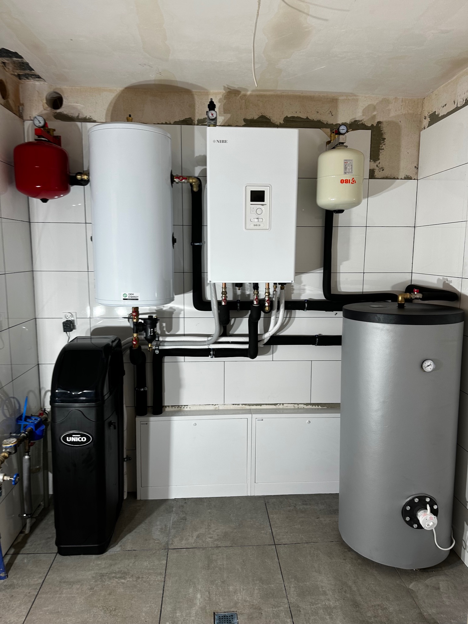 Kompletna instalacja grzewcza w kotłowni: piec gazowy Nibe, zbiorniki buforowe (czerwony i biały), szary zasobnik CWU i czarny zmiękczacz wody Unico. Białe płytki na ścianach.
