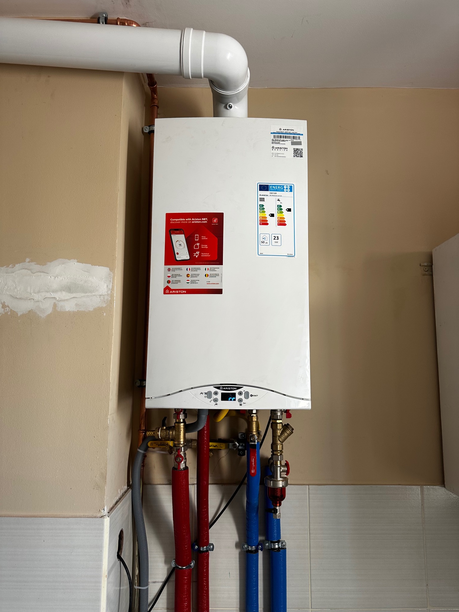 Biały piec gazowy Ariston na ścianie, z widocznymi rurami doprowadzającymi wodę (czerwone i niebieskie) i odprowadzającymi spaliny. Widoczna etykieta energetyczna.
