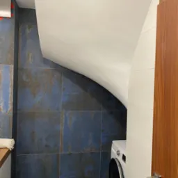 Niebieskie, kwadratowe płytki imitujące rdzę na ścianie łazienki, biały sufit ze skosem, fragment pralki i drewniane drzwi z chromowaną klamką.