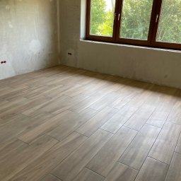 USŁUGI REMONTOWO - BUDOWLANE SŁAWOMIR CELIŃSKI-MYSŁAW - Wnętrze pokoju w trakcie wykańczania, z ułożoną podłogą z płytek imitujących drewno, surowe ściany z widocznymi puszkami elektrycznymi i oknem z widokiem na zieleń.