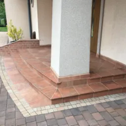 Wykończone płytkami schody wejściowe do domu w Wyrach, z kolumną i brukowanym chodnikiem. Widoczny fragment elewacji i drzwi.