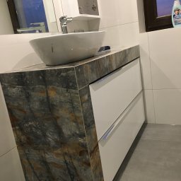 USŁUGI REMONTOWO - BUDOWLANE SŁAWOMIR CELIŃSKI-MYSŁAW - Nowoczesna umywalka nablatowa o owalnym kształcie, osadzona na blacie z płytek imitujących marmur, pod którą znajdują się białe szuflady z minimalistycznymi uchwytami, całość w jasnej łazience...