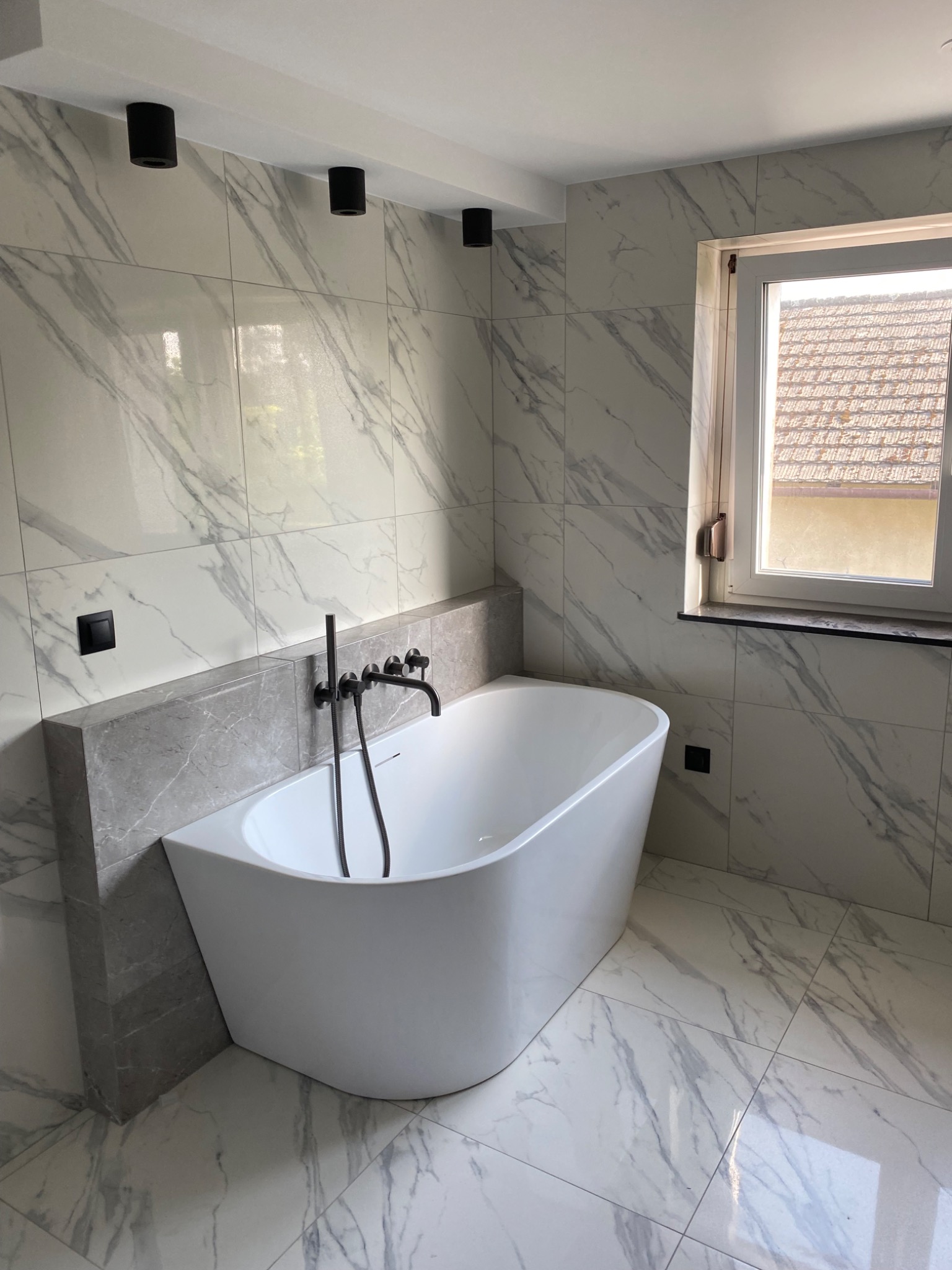 Nowoczesna łazienka z wolnostojącą białą wanną, armaturą w kolorze czarnym mat oraz marmurowymi płytkami na ścianach i podłodze, oświetlenie sufitowe punktowe.
