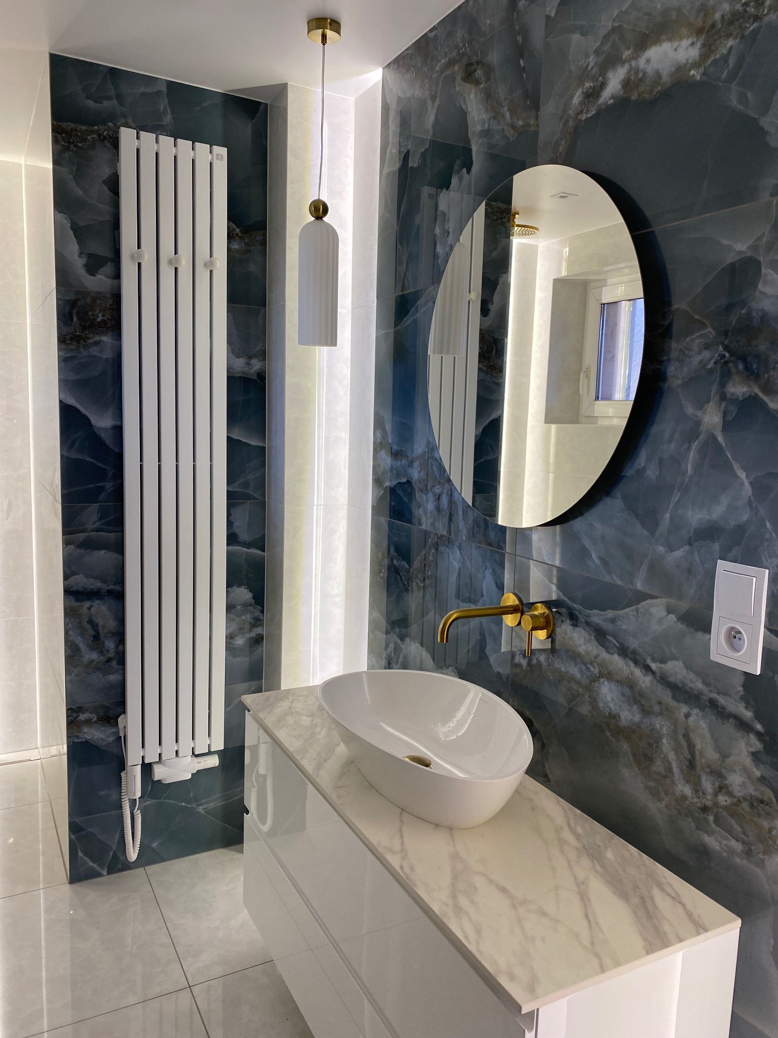 Nowoczesna łazienka z marmurowymi płytkami w odcieniach granatu, białym grzejnikiem drabinkowym, owalną umywalką nablatową z baterią w kolorze złotym oraz okrągłym lustrem z czarną ramą.