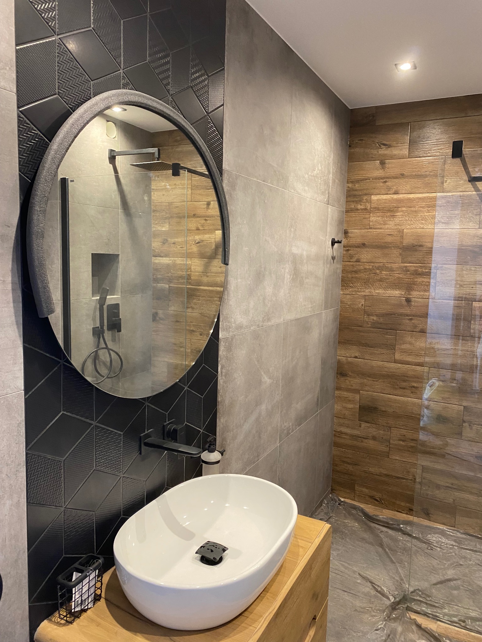Nowoczesna łazienka po remoncie z owalnym lustrem, umywalką nablatową na drewnianym blacie, czarną armaturą i geometrycznymi płytkami na ścianie.