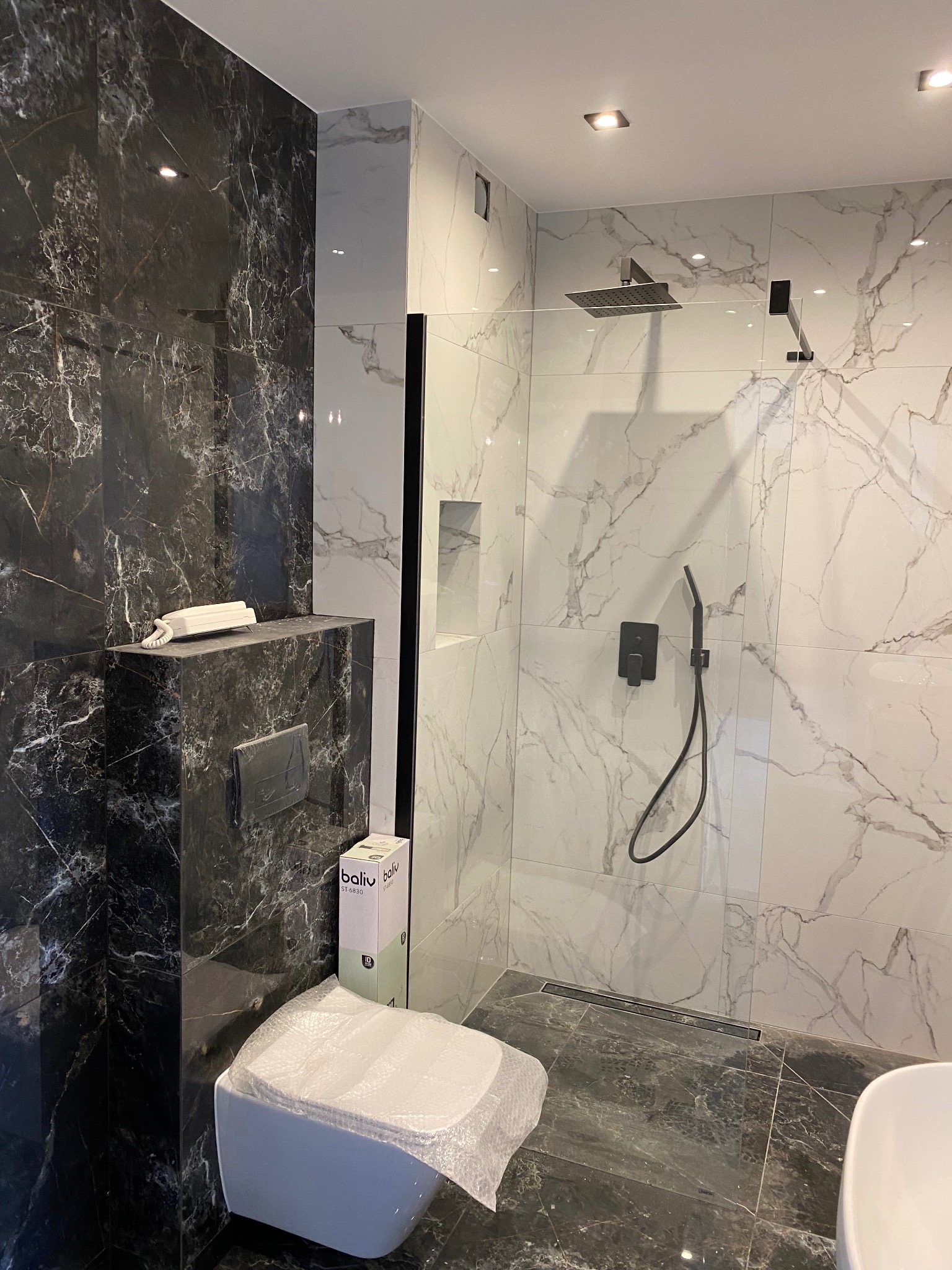 Nowoczesna łazienka z czarno-białymi marmurkowymi płytkami, deszczownicą w czarnym kolorze oraz wiszącą toaletą z opakowaniem producenta, częściowo jeszcze zabezpieczoną folią.