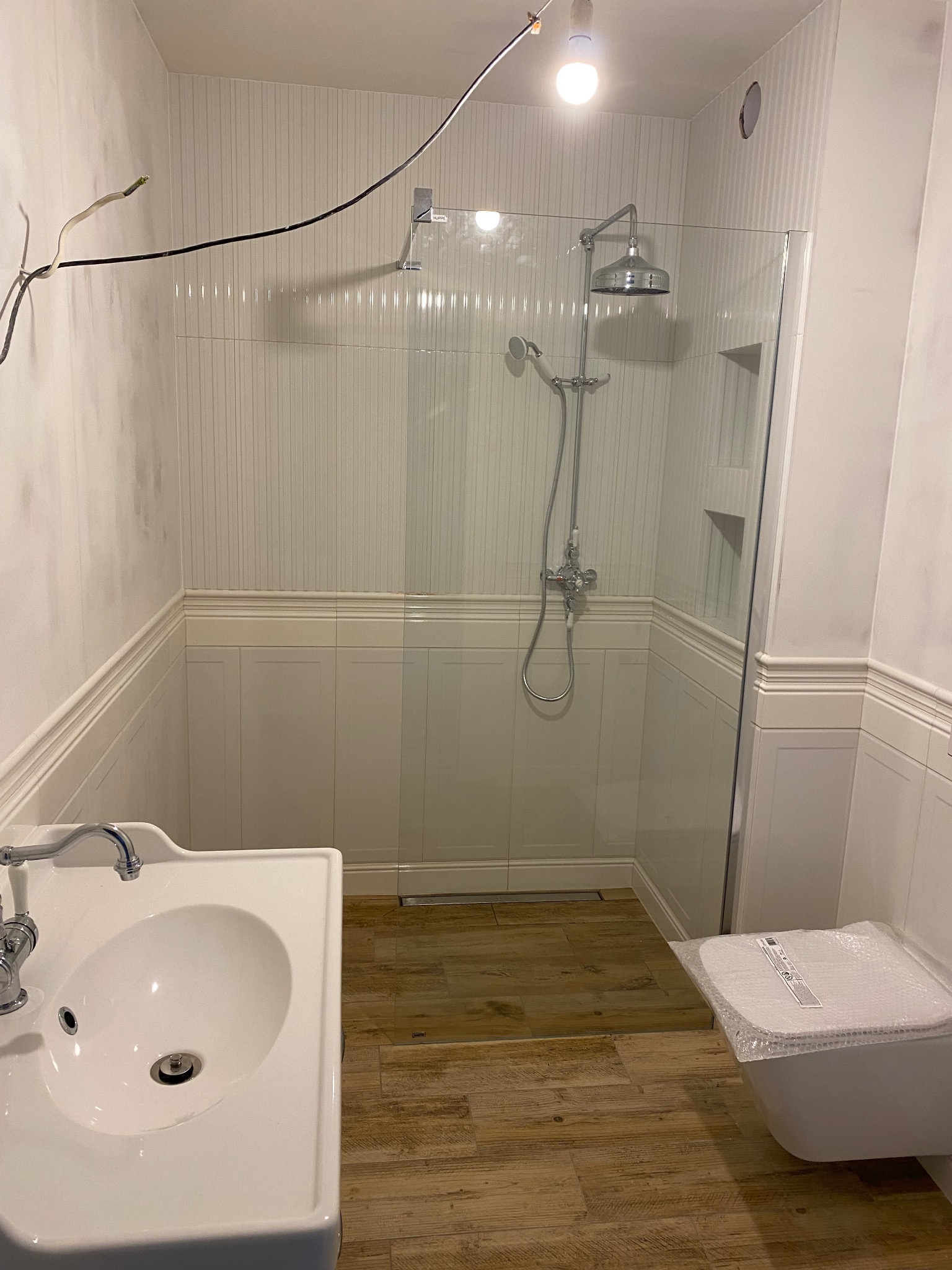 Nowoczesna łazienka w trakcie wykańczania: kabina prysznicowa ze szklaną ścianką, biała umywalka z chromowaną baterią i toaleta zabezpieczona folią, drewnopodobna podłoga, białe panele ścienne...