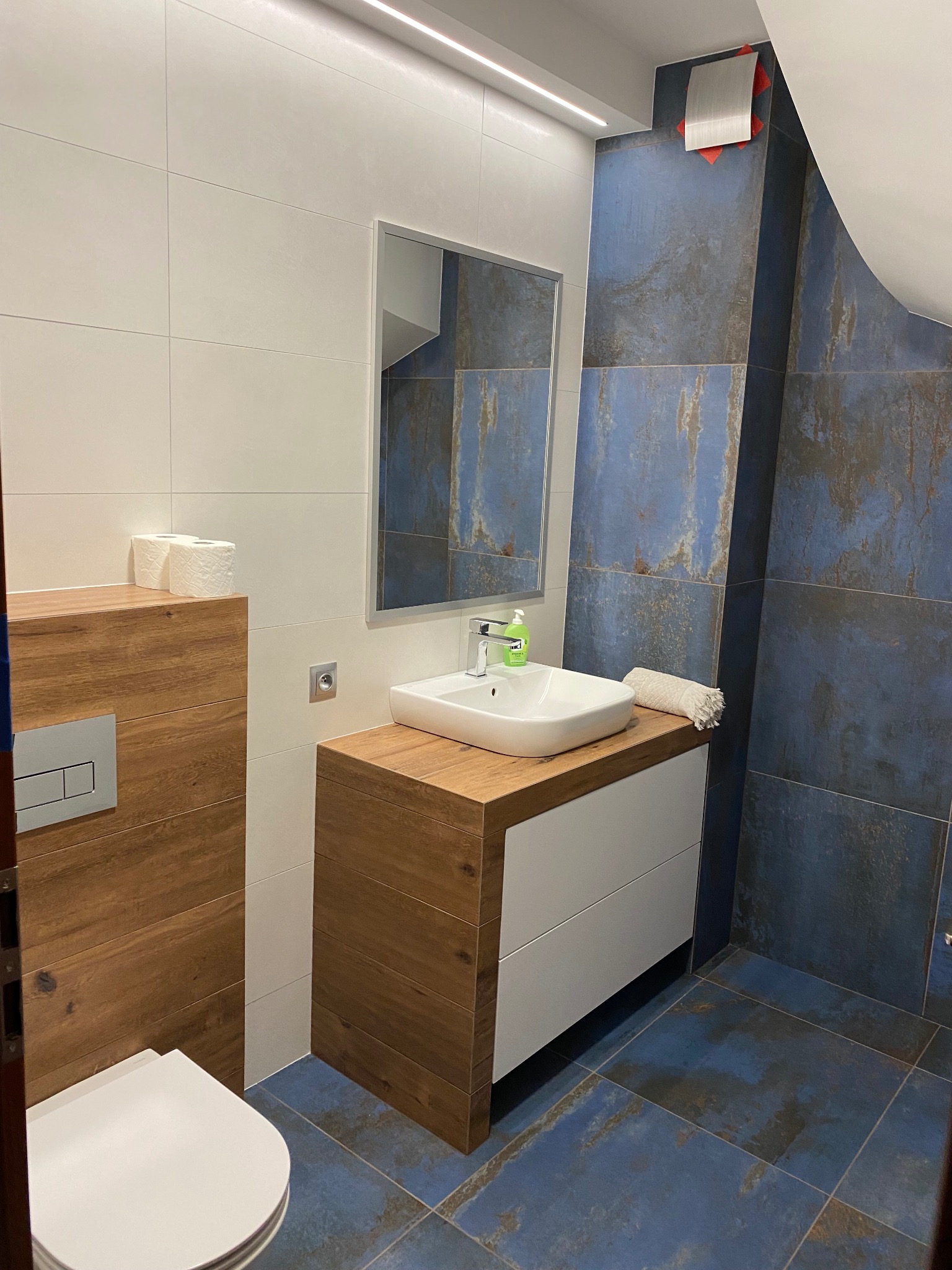 Nowoczesna łazienka z biało-niebieskimi płytkami imitującymi rdzę, drewnianą zabudową toalety i szafką pod umywalkę z drewnianym blatem, oświetleniem LED i prostokątnym lustrem.
