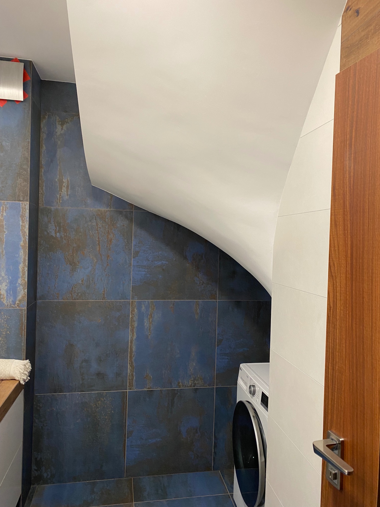 Niebieskie, kwadratowe płytki imitujące rdzę na ścianie łazienki, biały sufit ze skosem, fragment pralki i drewniane drzwi z chromowaną klamką.