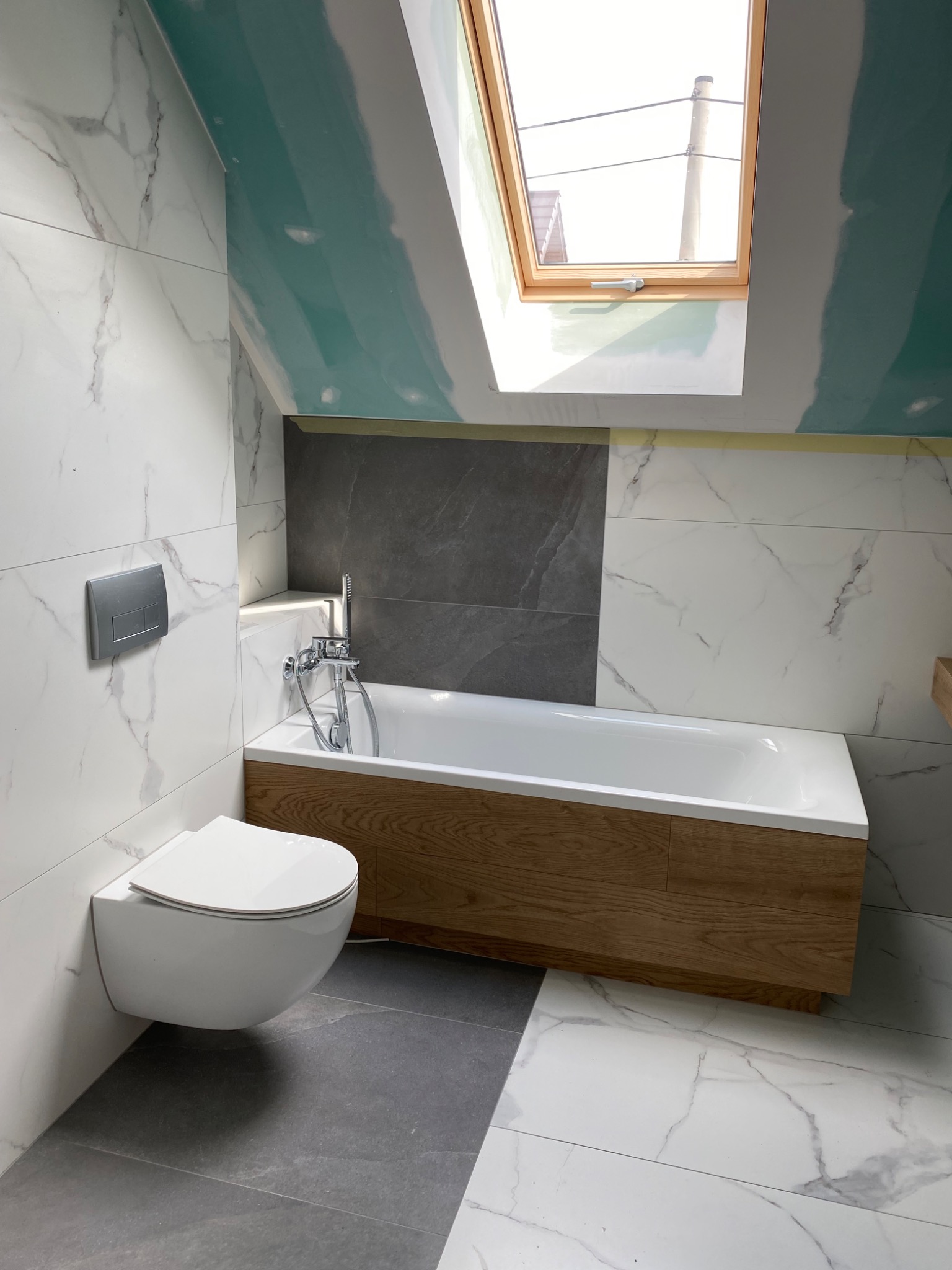 Nowoczesna łazienka w trakcie wykańczania: białe płytki imitujące marmur, szare płytki podłogowe, zabudowana wanna z drewnianym frontem, podwieszana toaleta, okno dachowe w skosie, widoczne ślady...