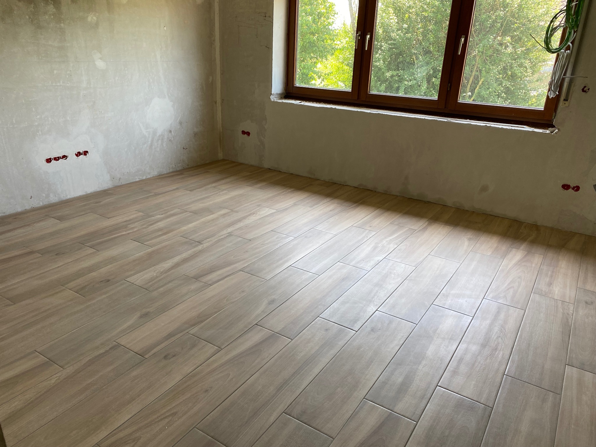 Wnętrze pokoju w trakcie wykańczania, z ułożoną podłogą z płytek imitujących drewno, surowe ściany z widocznymi puszkami elektrycznymi i oknem z widokiem na zieleń.