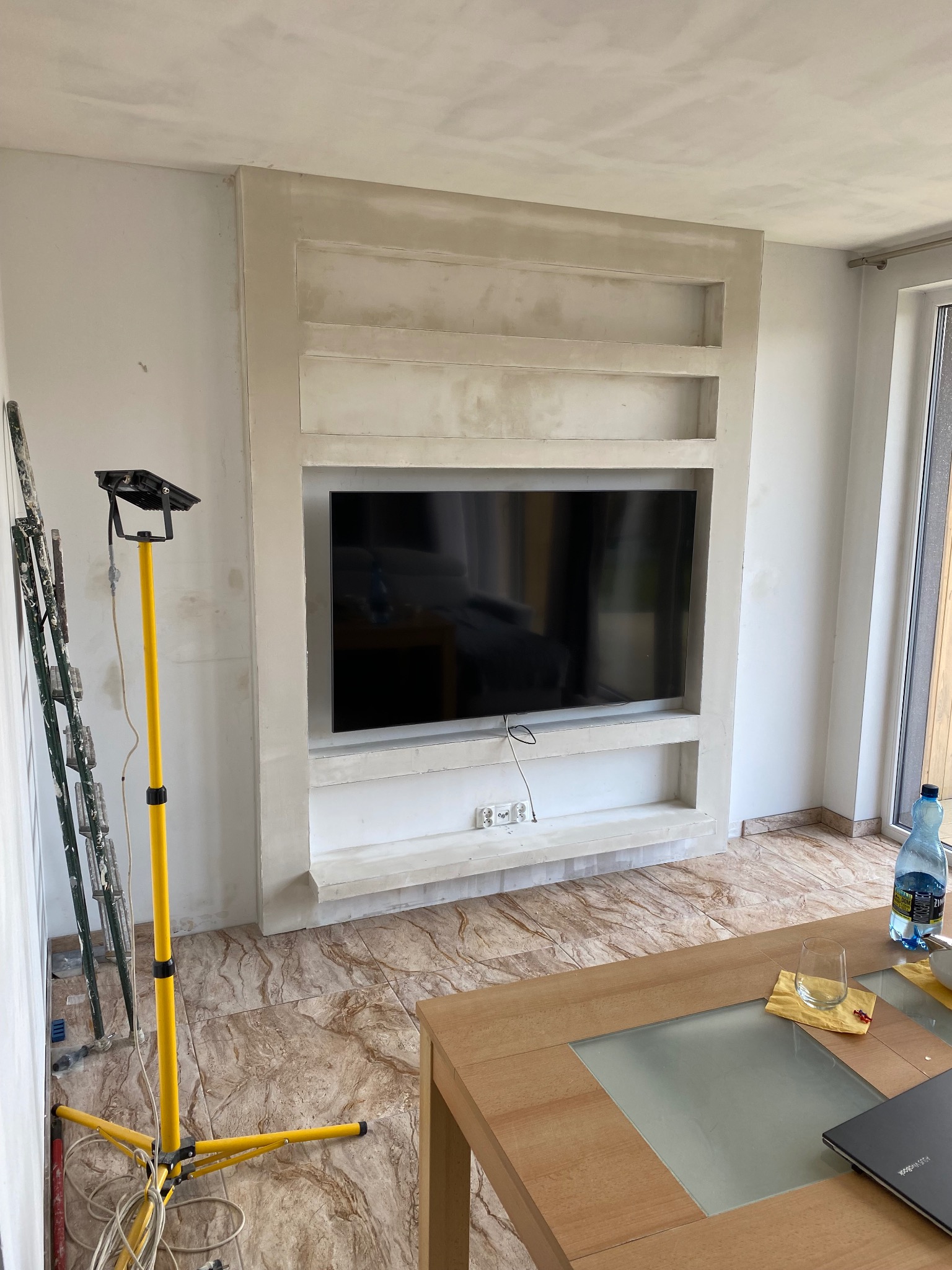 Ściana z płyt kartonowo-gipsowych w trakcie zabudowy pod telewizor, widoczne półki, lampa budowlana i drabina malarska, na podłodze płytki imitujące marmur.