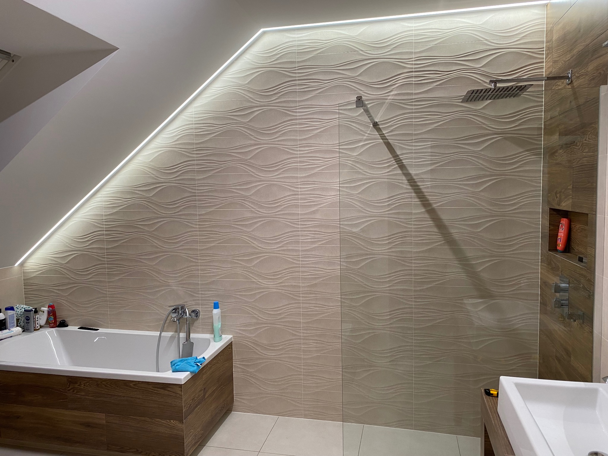 Nowoczesna łazienka na poddaszu z podświetlaną listwą LED przy skośnym suficie, beżowe płytki 3D imitujące fale, drewnopodobna obudowa wanny i kabina prysznicowa typu walk-in z deszczownicą.