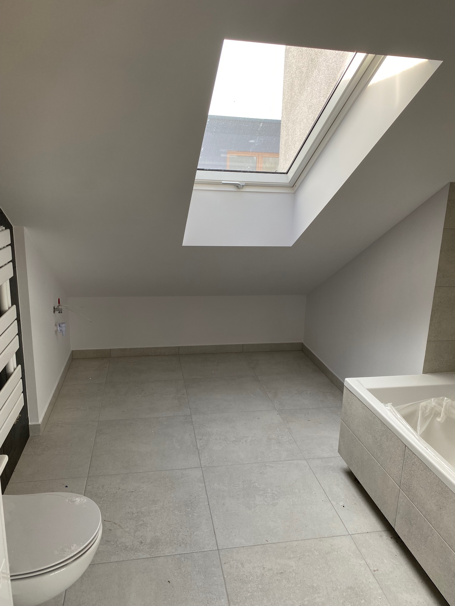Surowa łazienka na poddaszu z oknem dachowym, szarą glazurą podłogową i częściowo wykończoną obudową wanny oraz grzejnikiem drabinkowym. Widoczne przyłącza wodne i kanalizacyjne w ścianie.