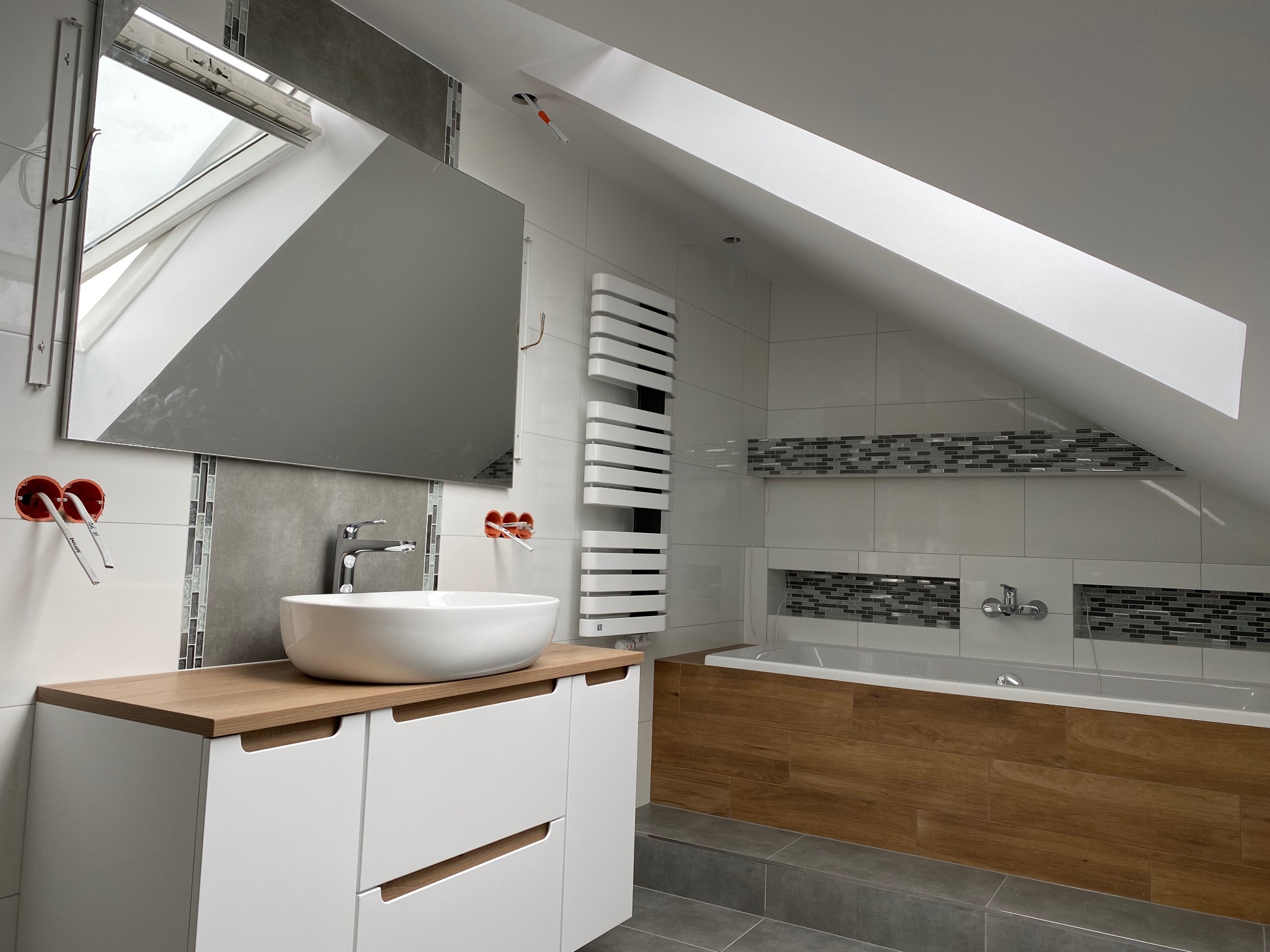 Nowoczesna łazienka na poddaszu z umywalką nablatową, drewnianym blatem, białą szafką pod umywalką i grzejnikiem drabinkowym, płytki imitujące drewno na obudowie wanny, widoczne przygotowane...