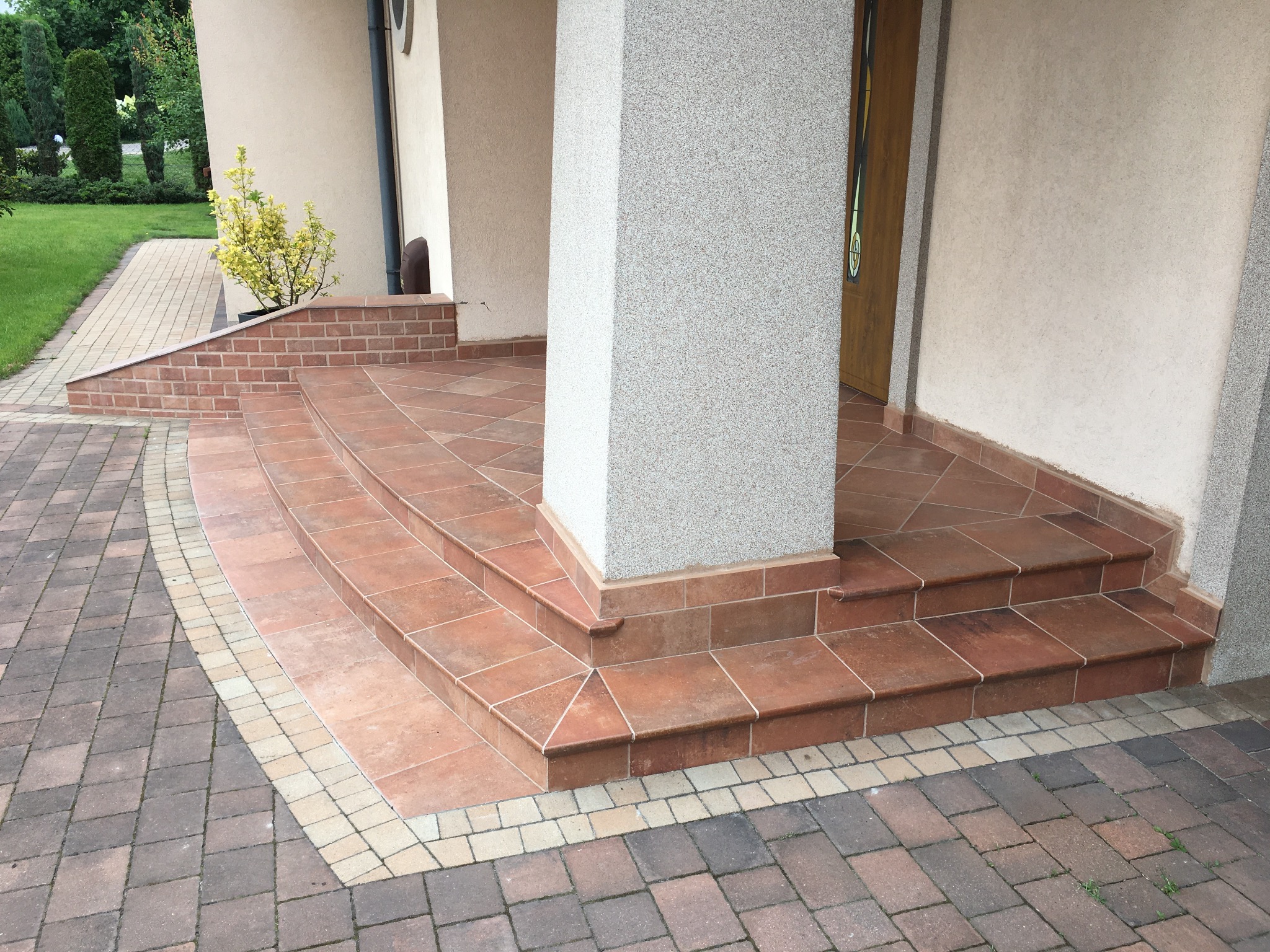 Wykończone płytkami schody wejściowe do domu w Wyrach, z kolumną i brukowanym chodnikiem. Widoczny fragment elewacji i drzwi.