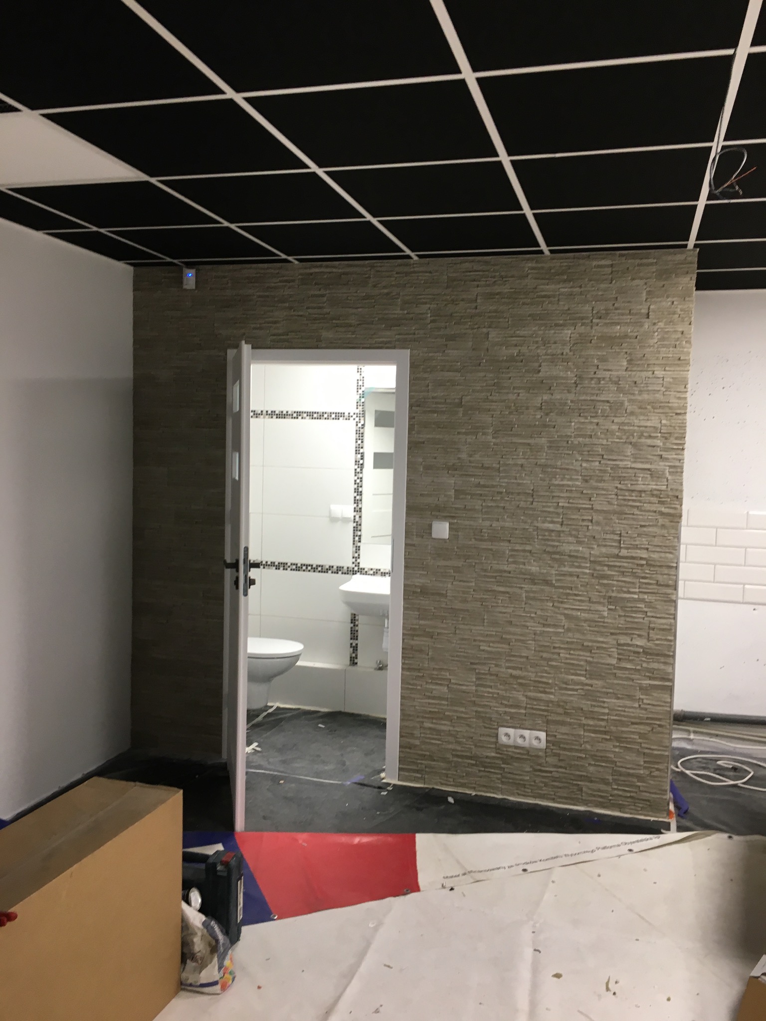 Fragment łazienki w trakcie prac wykończeniowych: ściana wyłożona dekoracyjnym kamieniem, widoczna toaleta i umywalka w głębi, czarny sufit kasetonowy i zabezpieczona folią podłoga.