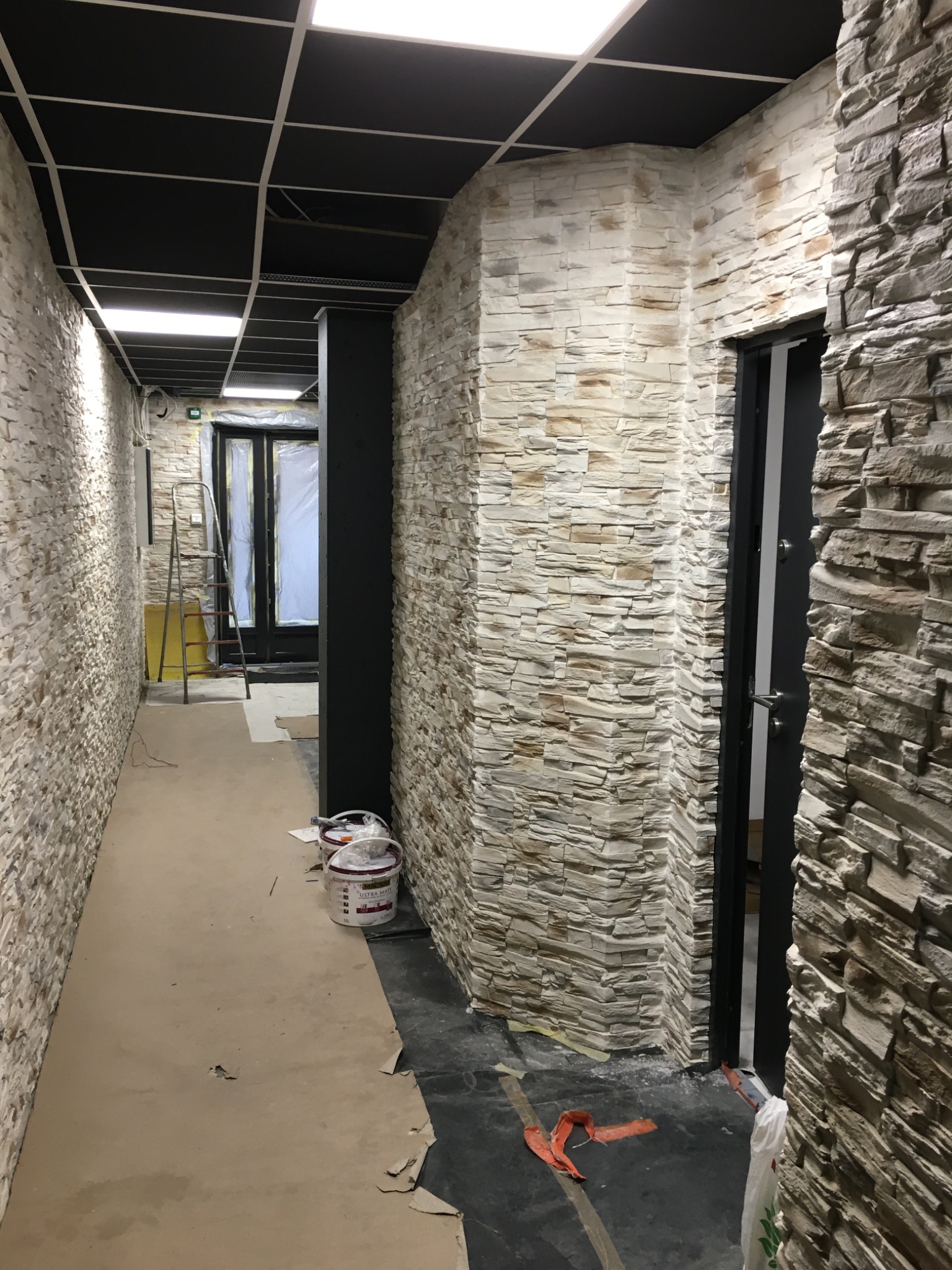 Korytarz w trakcie wykańczania z dekoracyjną okładziną kamienną na ścianach, widoczne puszki z farbą, zabezpieczona podłoga i drabina oparta o drzwi.