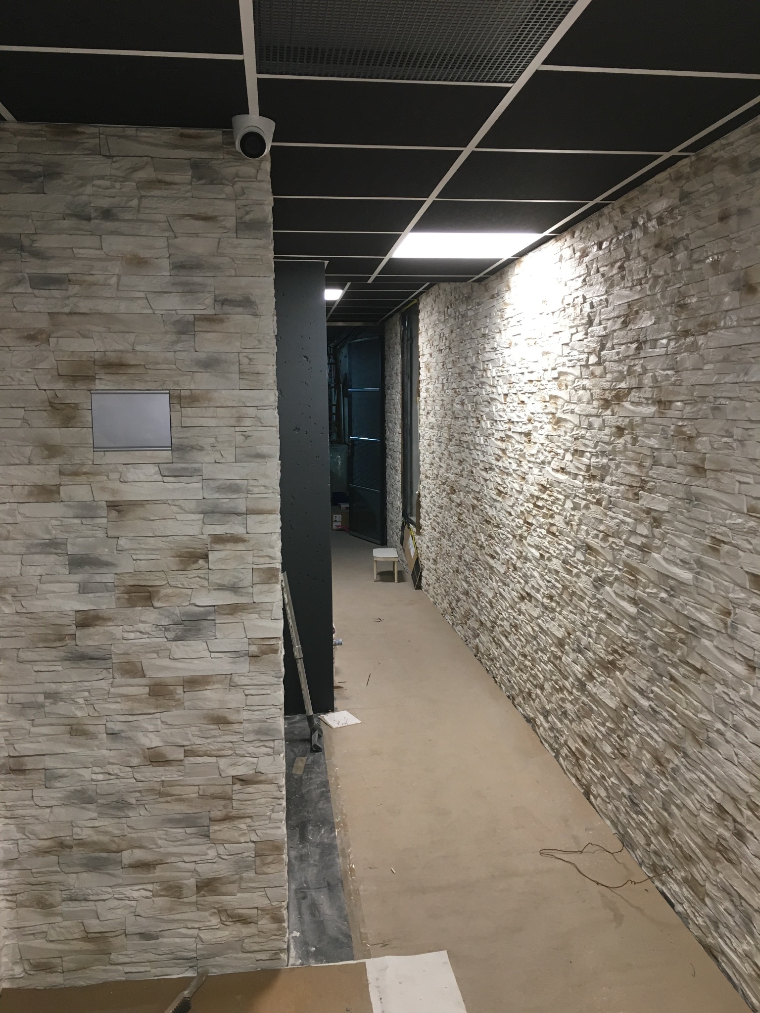 Korytarz w trakcie wykańczania z dekoracyjną okładziną kamienną na ścianach, czarnym sufitem rastrowym i widocznym fragmentem instalacji elektrycznej na podłodze.