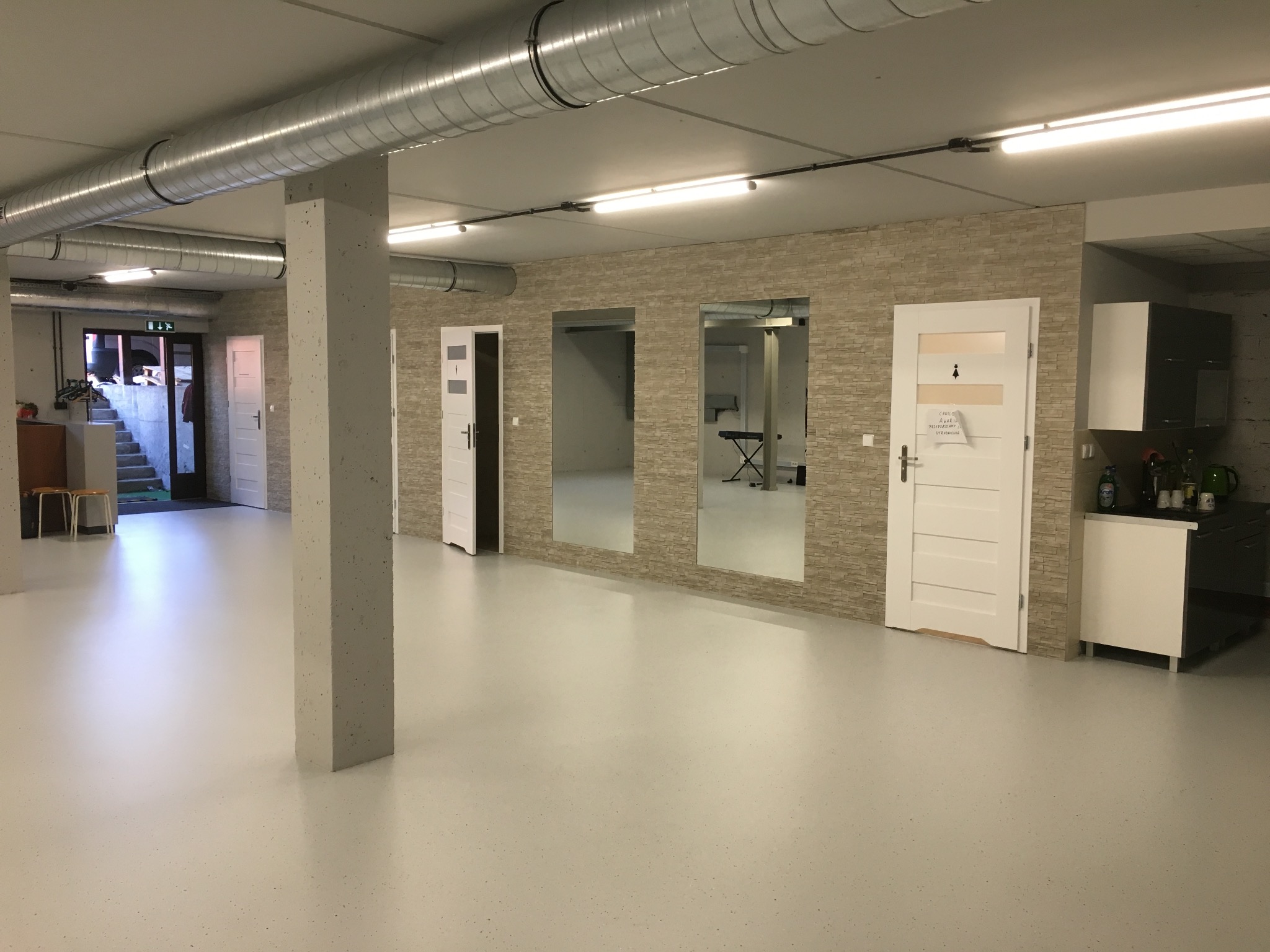 Przestronne, jasne wnętrze z szarą, żywiczną podłogą, ścianą wyłożoną dekoracyjną cegłą, białymi drzwiami i dużymi lustrami w ścianie, widoczne instalacje wentylacyjne pod sufitem, oświetlenie...