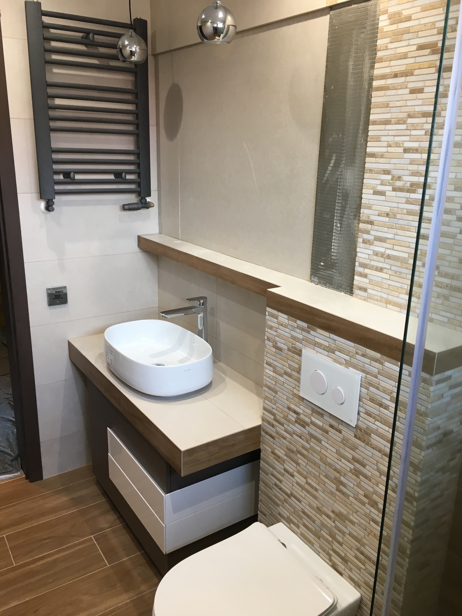 Nowoczesna łazienka w trakcie wykańczania, widoczna owalna umywalka nablatowa, grzejnik drabinkowy, częściowo ułożona mozaika ścienna oraz stelaż podtynkowy WC.