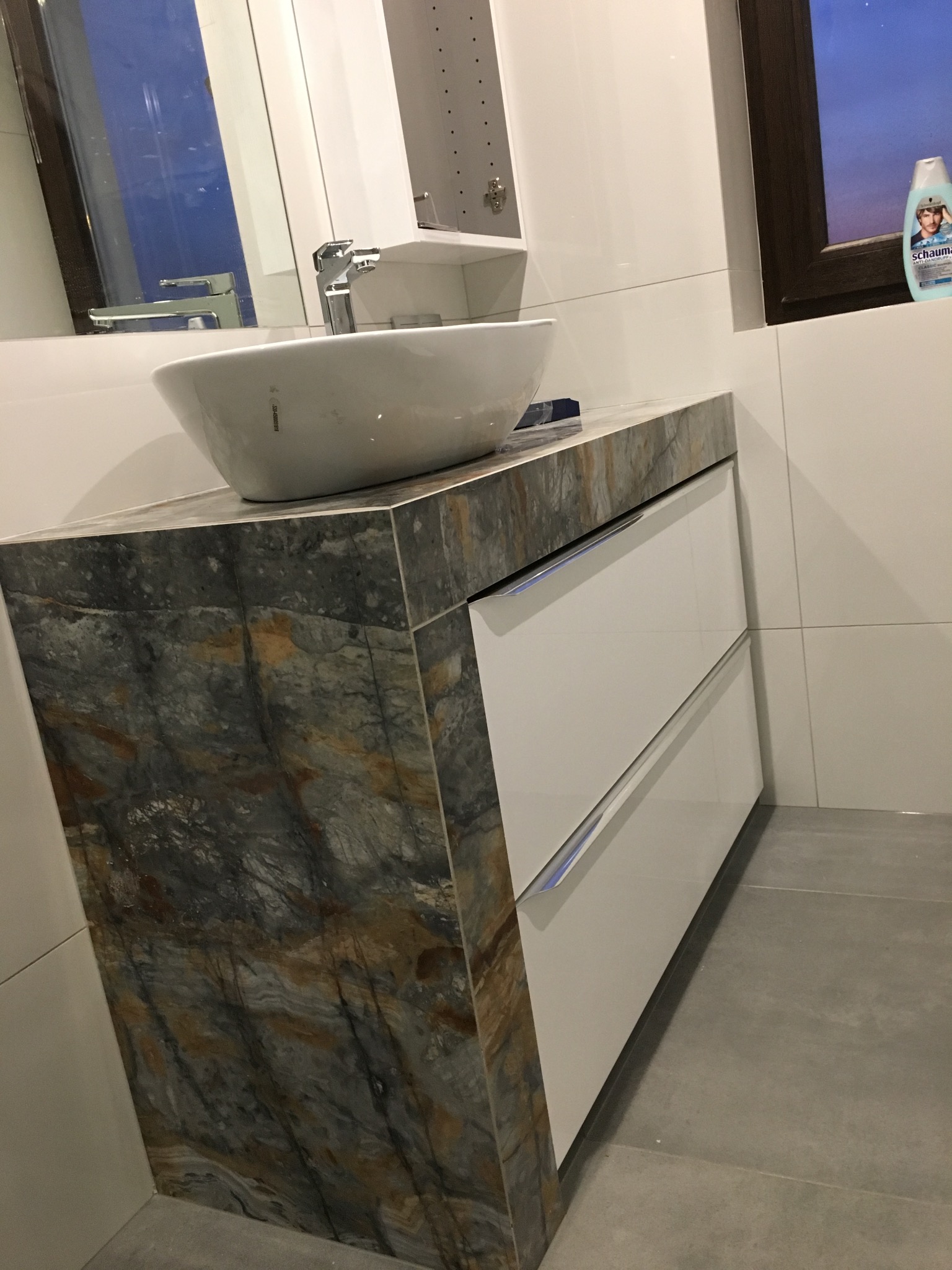 Nowoczesna umywalka nablatowa o owalnym kształcie, osadzona na blacie z płytek imitujących marmur, pod którą znajdują się białe szuflady z minimalistycznymi uchwytami, całość w jasnej łazience...