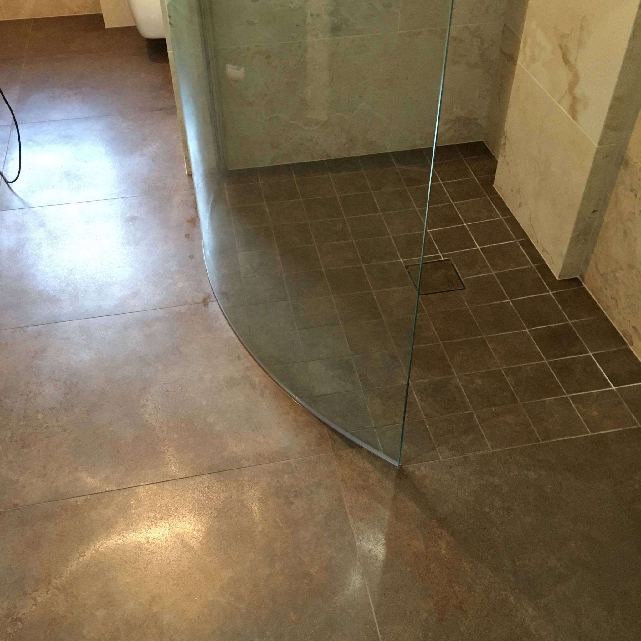 Nowoczesna kabina prysznicowa z zakrzywioną szybą, brązowe płytki podłogowe w dwóch odcieniach, beżowe płytki ścienne, widoczny fragment toalety w tle.