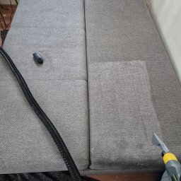 Pan Plama - Szara kanapa modułowa, częściowo wyczyszczona, z widoczną różnicą w odcieniu, dysza odkurzacza piorącego z żółtymi elementami w prawym dolnym rogu, obok czarny wąż i mała szczotka.