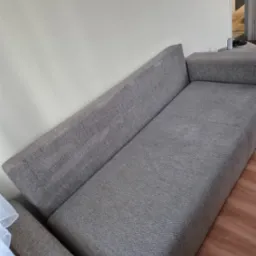 Szara kanapa z widocznym środkiem do czyszczenia tapicerki i szczotką na boku, ustawiona przy białej ścianie i drewnianej podłodze.