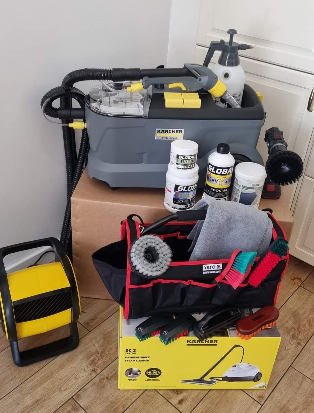 Profesjonalny sprzęt do czyszczenia tapicerki i dywanów marki Karcher, wraz z zestawem szczotek, środków czyszczących Global i torbą narzędziową Yato, ustawiony na kartonach na drewnianej podłodze.