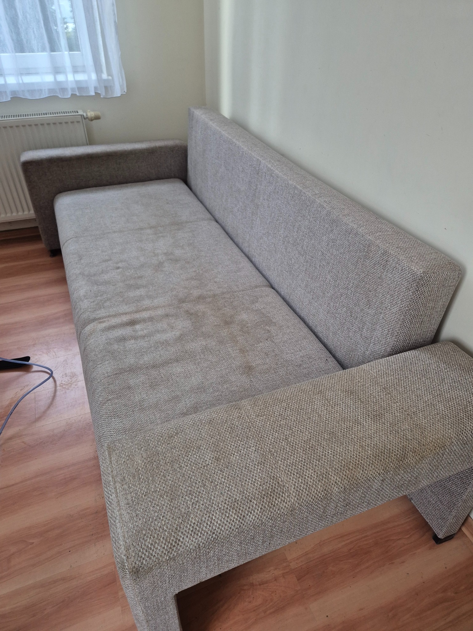 Beżowa, materiałowa kanapa z widocznymi plamami na siedzisku, stojąca na panelach podłogowych, z grzejnikiem i zasłoniętym oknem w tle.